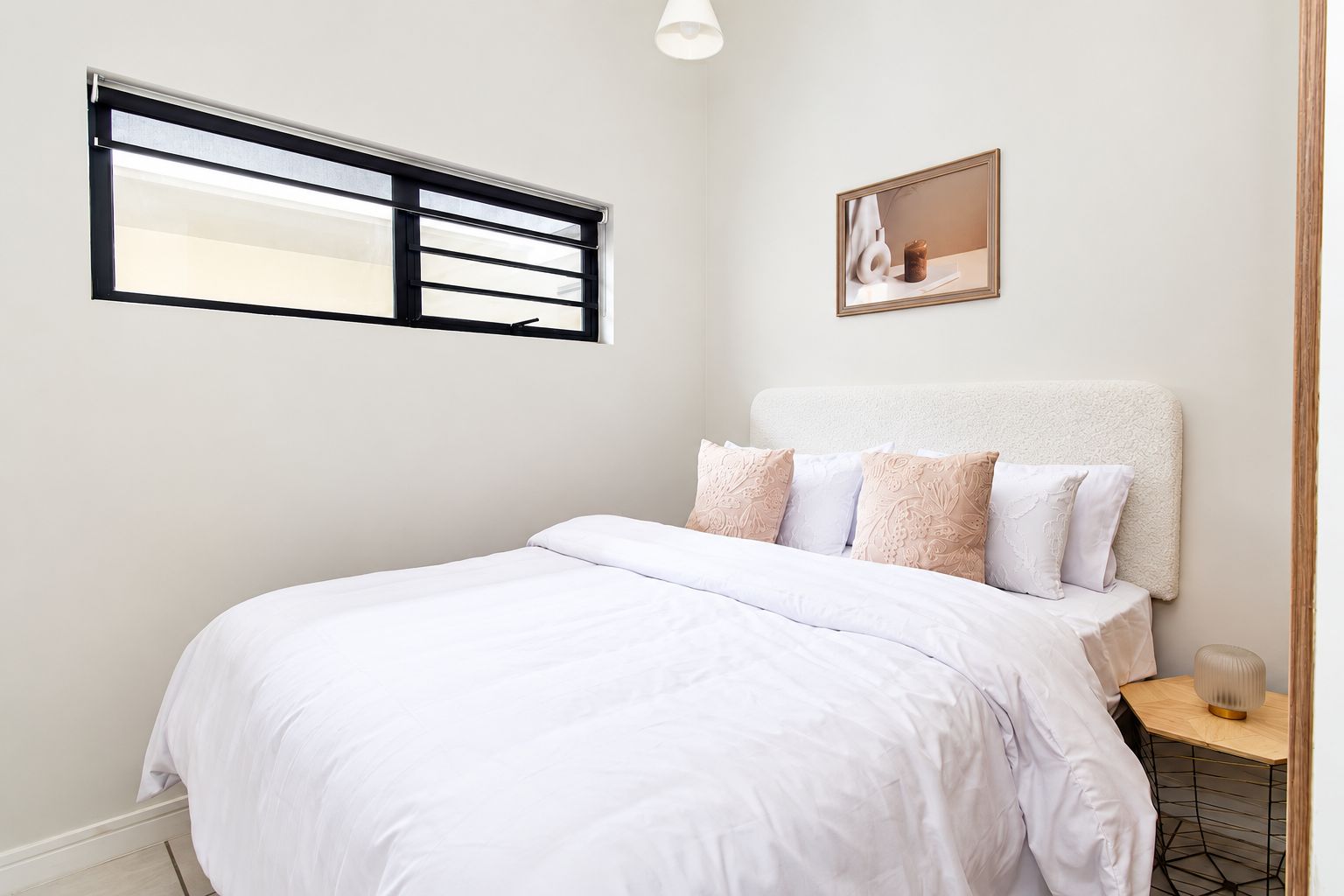 Une chambre à coucher avec un lit, une table de chevet, une lampe, une fenêtre avec des stores et un tableau encadré au mur.