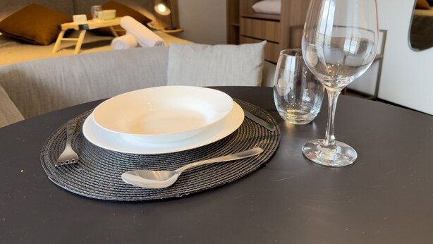 Una mesa de comedor dispuesta con un plato blanco, una copa, una cuchara y un tenedor, sobre un mantel tejido.