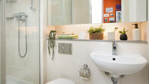 Casa de banho moderna com cabine de duche em vidro, lavatório branco e espelho que reflete um quarto com paredes laranja.