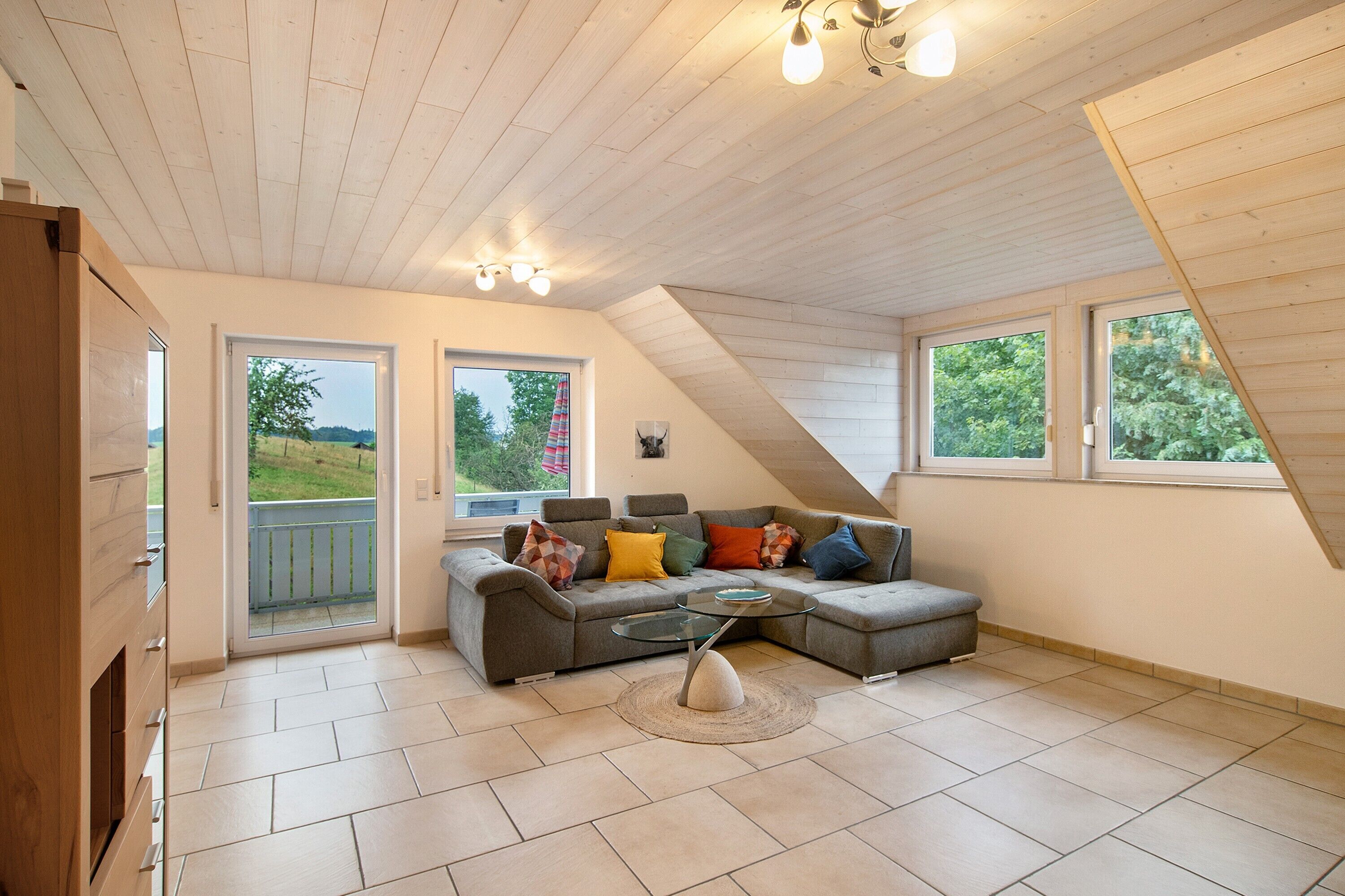 Une chambre moderne de style loft, dotée d’un canapé d’angle, d’une table basse en verre et offrant une vue sur un paysage verdoyant grâce à de grandes fenêtres.