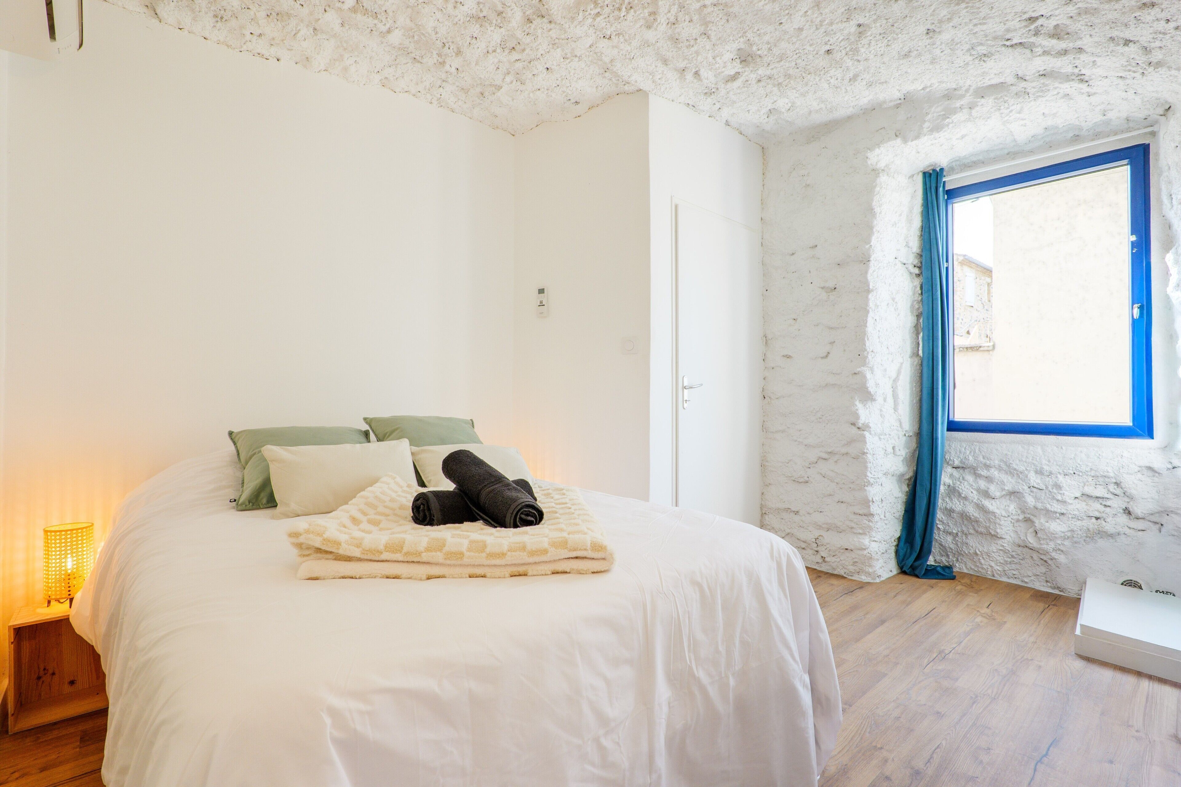 Une chambre à coucher avec un lit, des oreillers, une couverture, une table de chevet avec une lampe et une fenêtre avec des rideaux.