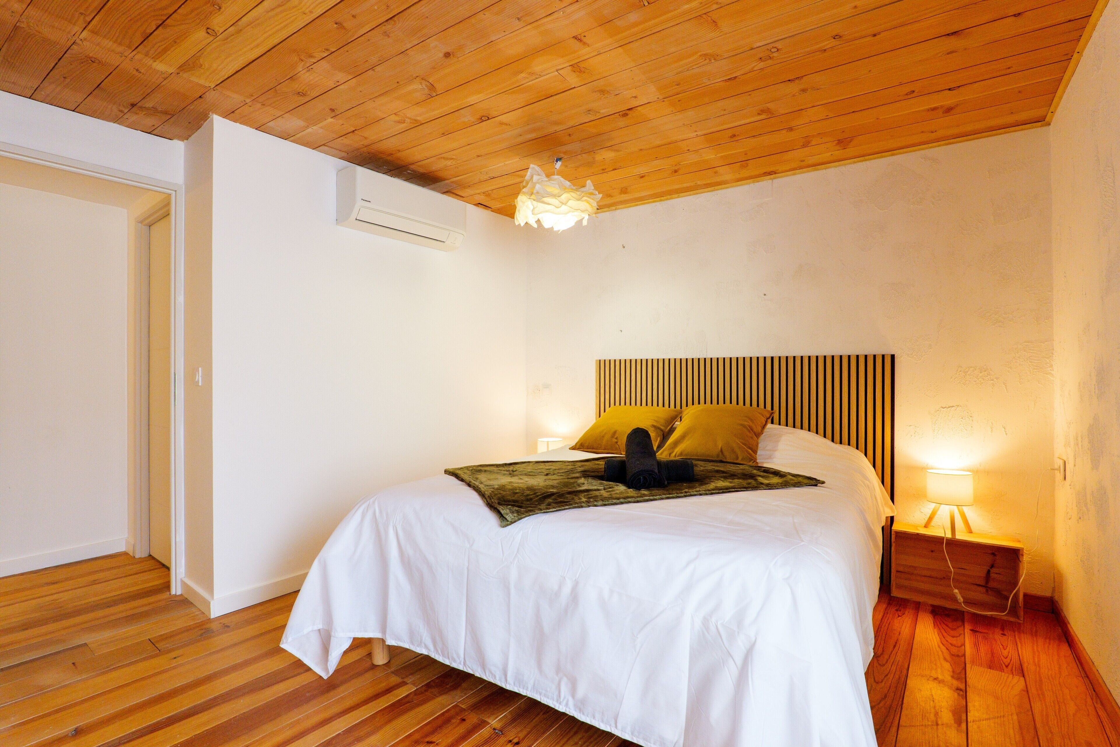 A bedroom with a wooden ceiling, a bed with white bedding, two pillows, a headboard with vertical slats, a bedside lamp, and an air conditioning unit.