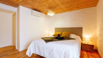 A bedroom with a wooden ceiling, a bed with white bedding, two pillows, a headboard with vertical slats, a bedside lamp, and an air conditioning unit.
