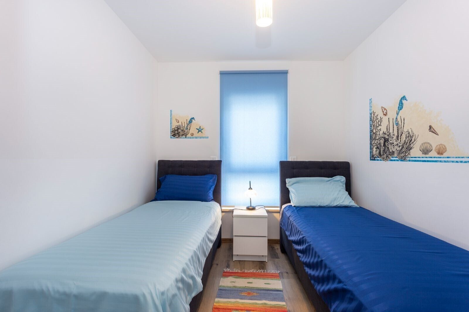 A bedroom with two single beds, a nightstand, a lamp, and a window with a blue blind.