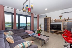 A modern living room with a sofa, coffee table, and TV, large windows with curtains, and a balcony with a view. - Premium beachfront apartment (Banjole)