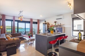 A modern living space with a dining area, kitchen, and large windows overlooking a body of water. - Premium beachfront apartment (Banjole)