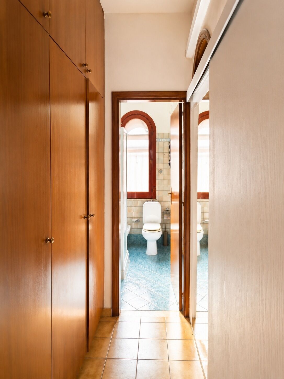 Een gang die leidt naar een badkamer met toilet, betegelde vloer en een houten garderobe.