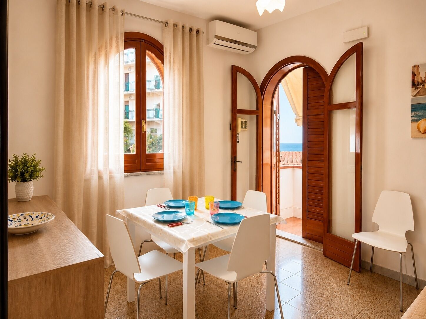 A dining area with a table set for two, white chairs, a potted plant, and a view of balconies and the sea.