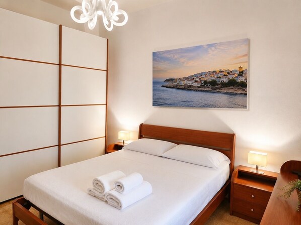 Ein ordentlich bezogenes Bett mit aufgerollten Handtüchern, einer Nachttischlampe und einem Bild einer Küstenstadt an der Wand.