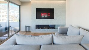 Ett modernt vardagsrum med en öppen spis, en väggmonterad tv som visar ”NETFLIX”, och ett stort fönster med utsikt över byggnader.