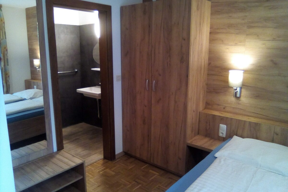 A bedroom with a wooden wardrobe, a bedside table, a mirror, and a wall-mounted light fixture.