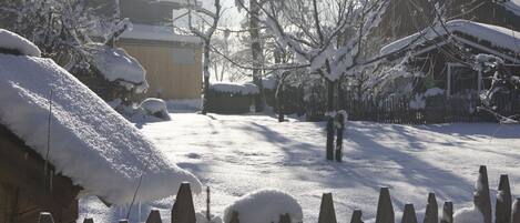 A snow-covered yard with a wooden fence, a building with a snow-covered roof, and bare trees.