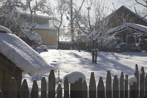 A snow-covered yard with a wooden fence, a building with a snow-covered roof, and bare trees.