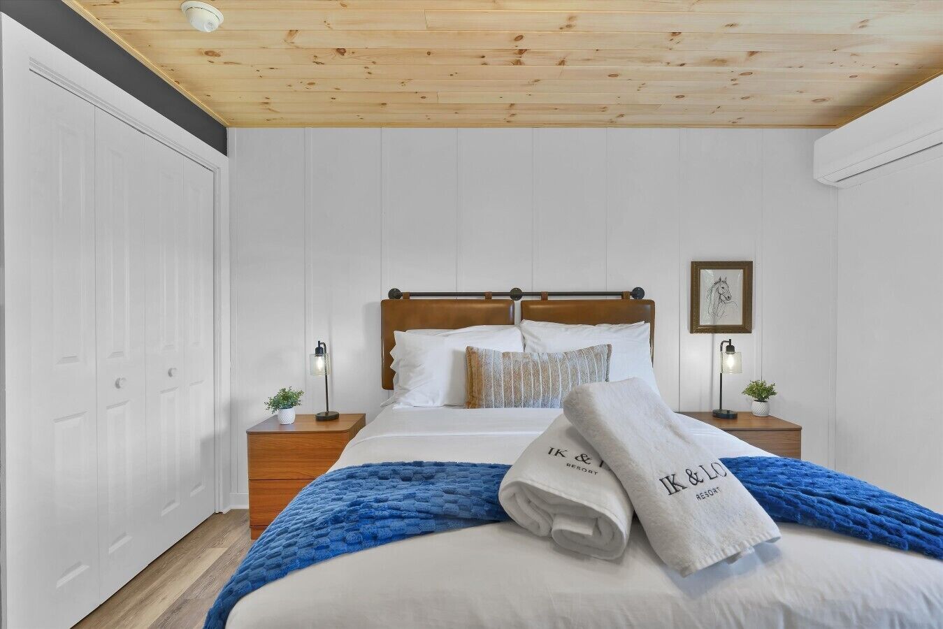 A bedroom with a wooden ceiling, a bed with white bedding, two bedside tables with lamps, and a framed picture on the wall.