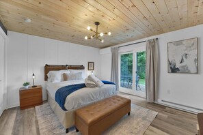 Un dormitorio con techo de madera, una cama con cabecera, un banco, una mesita de noche y una ventana con vistas a zonas verdes.
