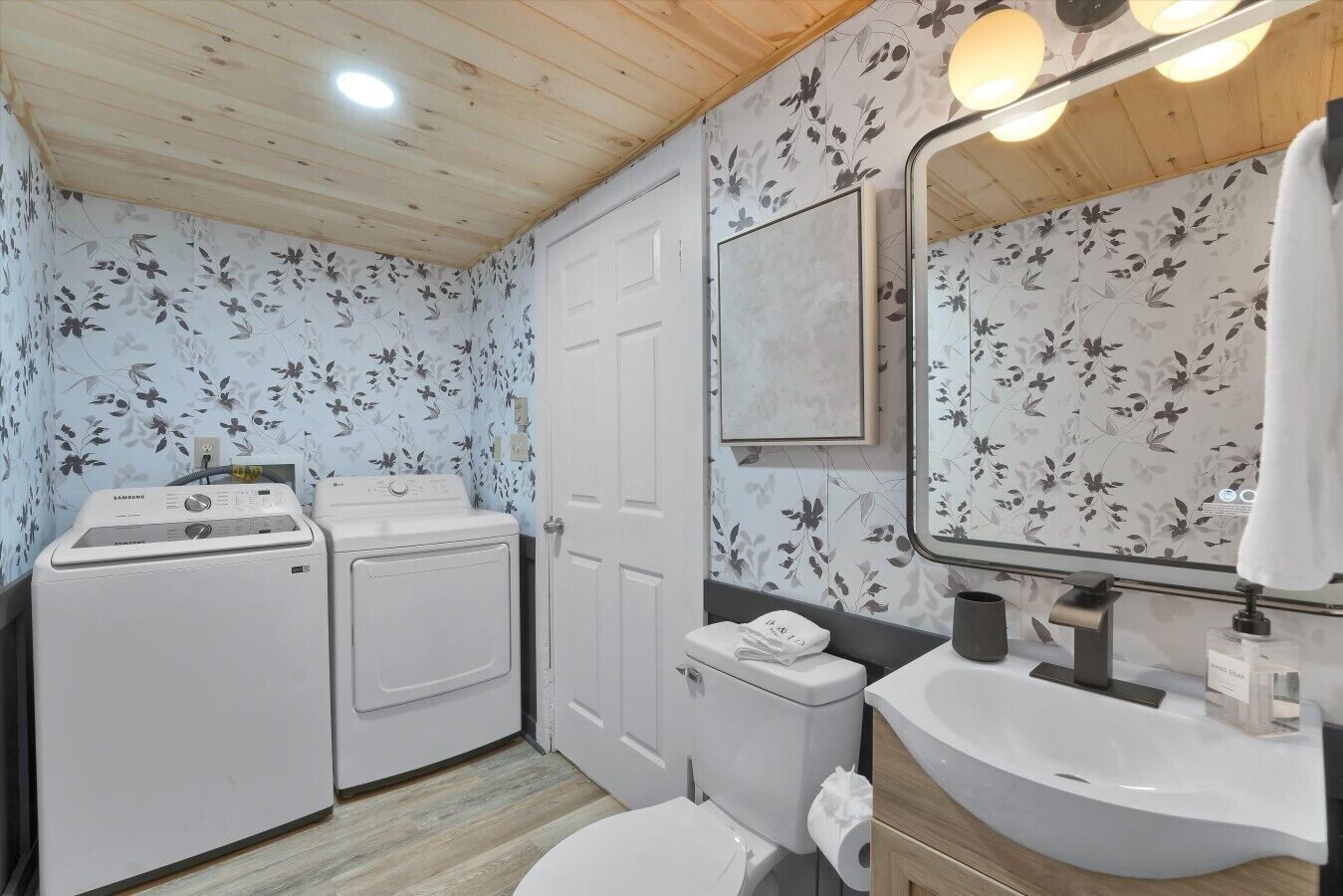 A compact bathroom with a washing machine, toilet, sink, and mirror.