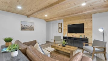 Una sala de estar con techo de madera, un televisor de pantalla plana montado en la pared, un sofá de cuero marrón, una mesa de centro de madera y un sillón gris.
