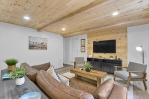 Ein Wohnzimmer mit Holzboden, einem Wand-Fernseher, einem braunen Ledersofa, einem Holztisch und einem grauen Sessel.