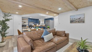 A modern living room with a brown leather sofa, a dining area, and a kitchen with blue cabinets.