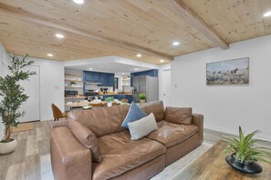 A modern living room with a brown leather sofa, a dining area, and a kitchen with blue cabinets.