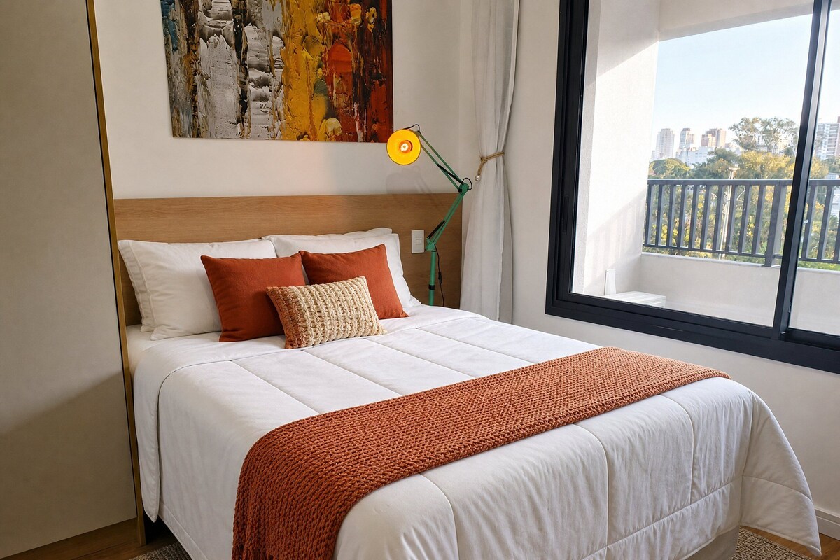 A neatly made bed with a white duvet and orange accents, a modern lamp, and a window with a view of buildings and trees.