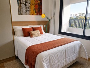 A neatly made bed with a white duvet and orange accents, a modern lamp, and a window with a view of buildings and trees. - Chanes Street (São Paulo)