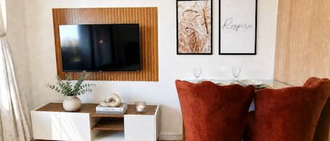 A modern living room with a flat-screen TV mounted on a wooden panel, a white console table with books and a vase, two framed artworks, and a pair of red velvet chairs.