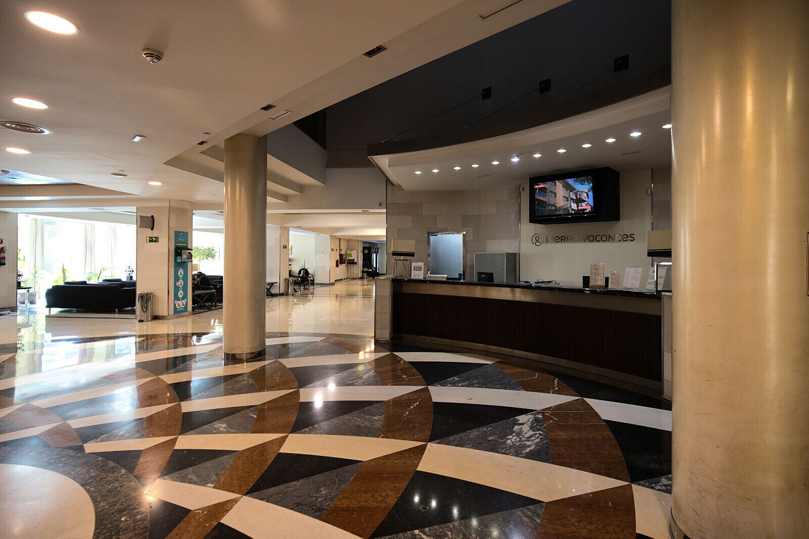 Vestíbulo de hotel con mostrador de recepción, un escritorio de información y una pantalla grande que muestra un programa.