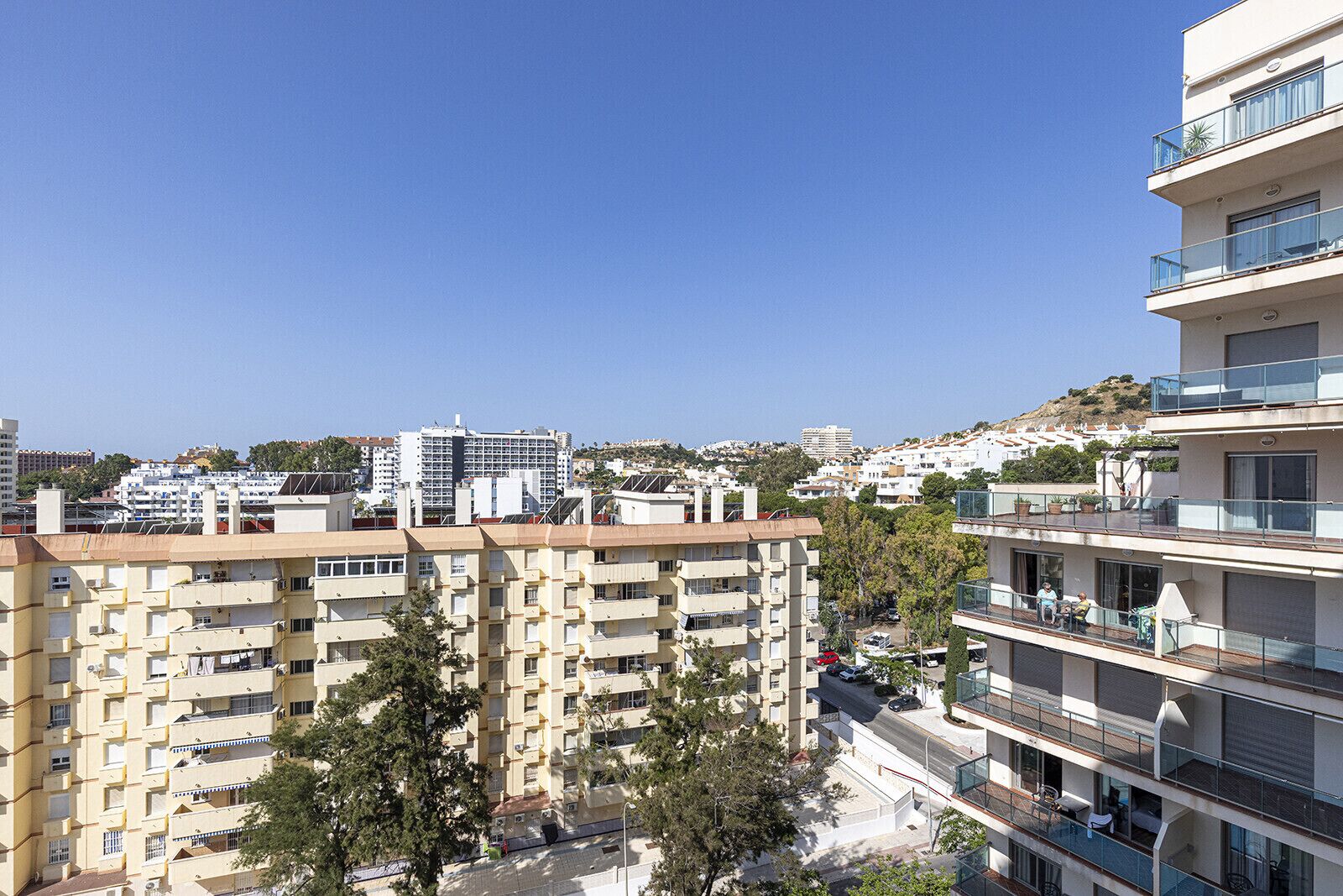 A multi-story residential building with balconies, a clear sky, and a hilly landscape in the background.