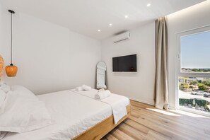 A bedroom with a bed, a TV, an air conditioner, a window with a view, and a hanging light fixture. - Aqua Breeze Golem 2BR Apartment (Golem)