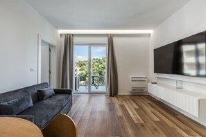 A modern living room with a sofa, a flat-screen TV, a sliding glass door to a balcony, and a white cabinet. - Aqua Breeze Golem 2BR Apartment (Golem)