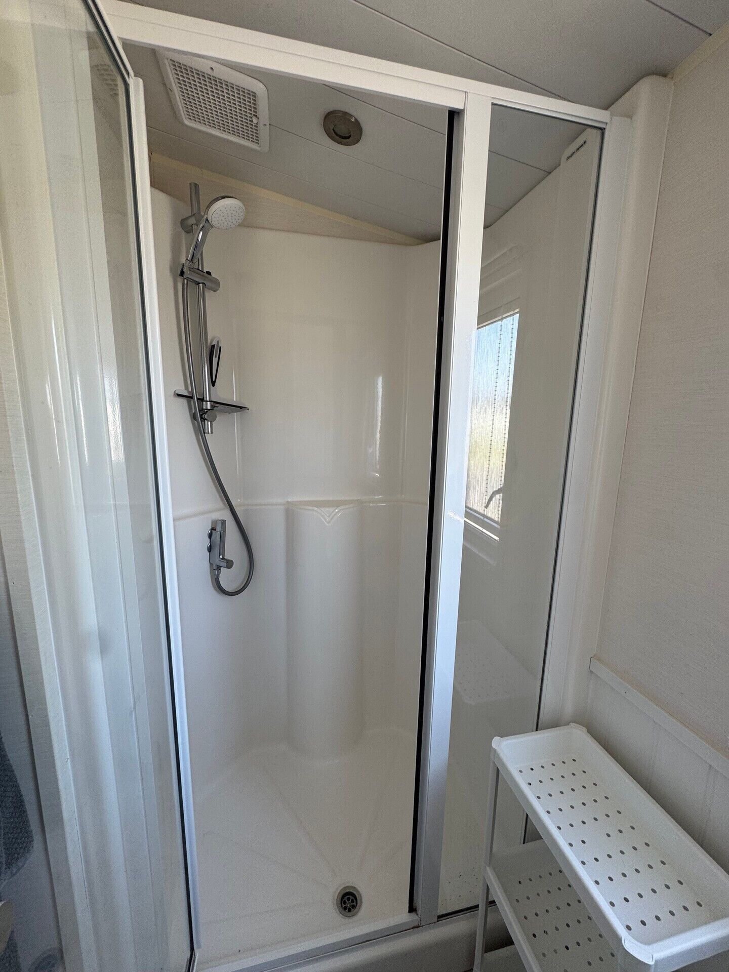 A glass-enclosed shower area with a handheld showerhead, a wall-mounted faucet, and a small shelf with a perforated tray.