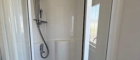 A glass-enclosed shower area with a handheld showerhead, a wall-mounted faucet, and a small shelf with a perforated tray.