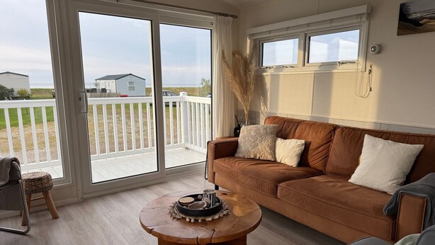 A living room with a brown leather sofa, a wooden coffee table, and a view through large sliding glass doors to a fenced outdoor area. - Camping Zeezicht - Chalet 16 (Westerland)