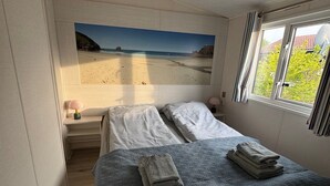 A bedroom with a bed, a nightstand, a window with curtains, and a beach view painting on the wall. - Camping Zeezicht - Chalet 16 (Westerland)