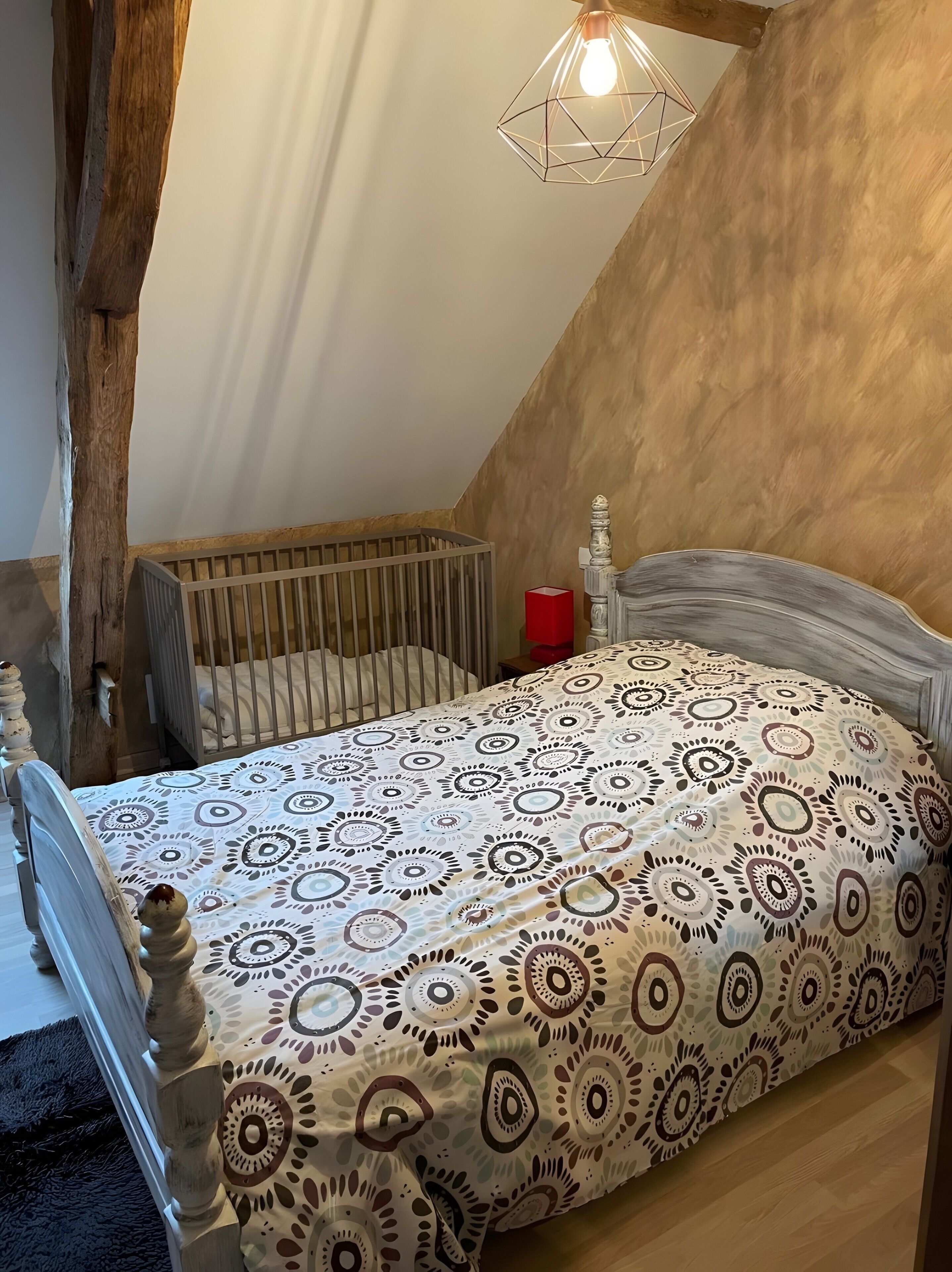 Ein Schlafzimmer mit schrägem Dach, einem Babybett, einem Bett mit gemusterten Betttextilien, einem dekorativen Kopfteil und einer hängenden Leuchte.