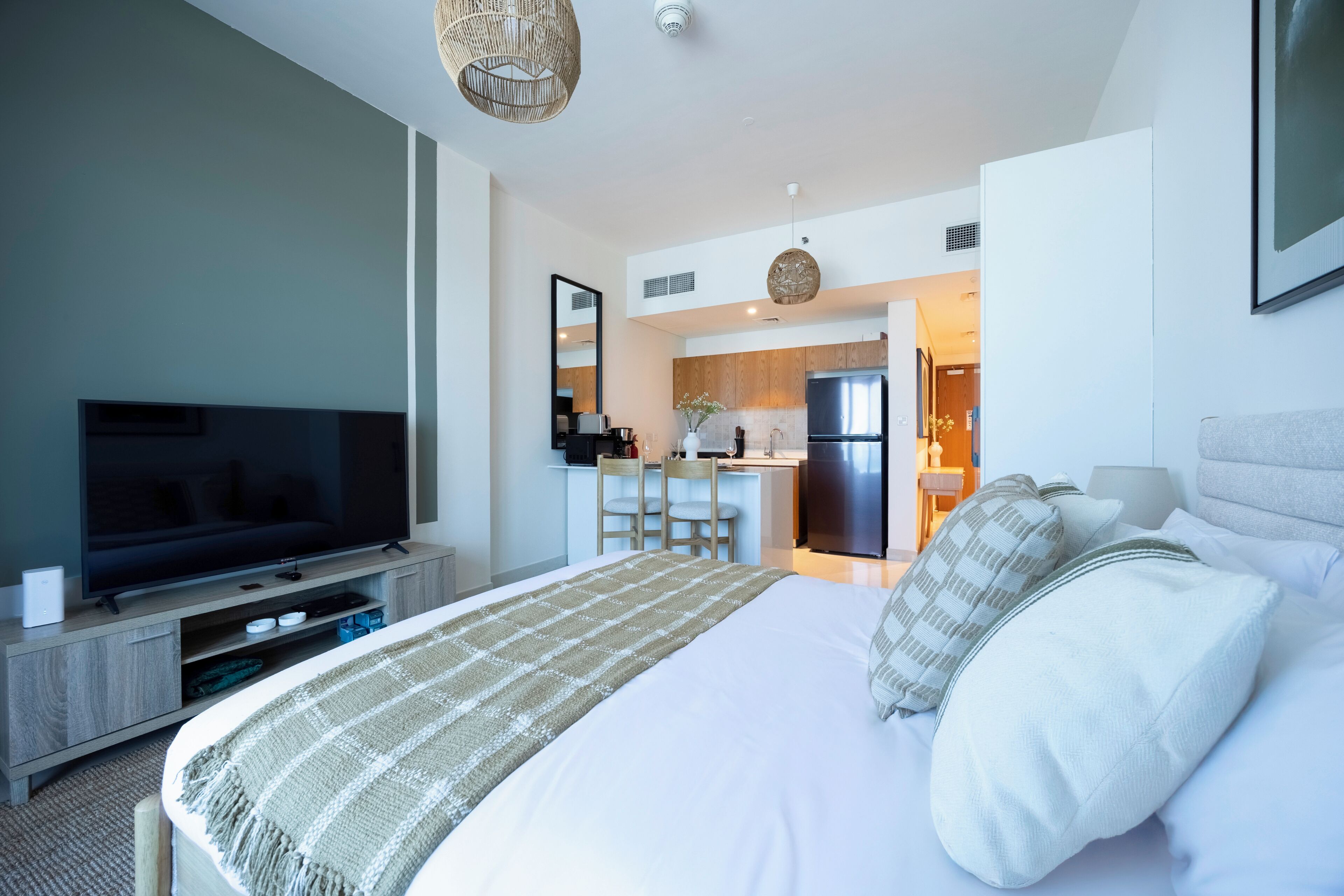 A modern hotel room with a large bed, a flat-screen TV, a kitchenette, and a dining area.