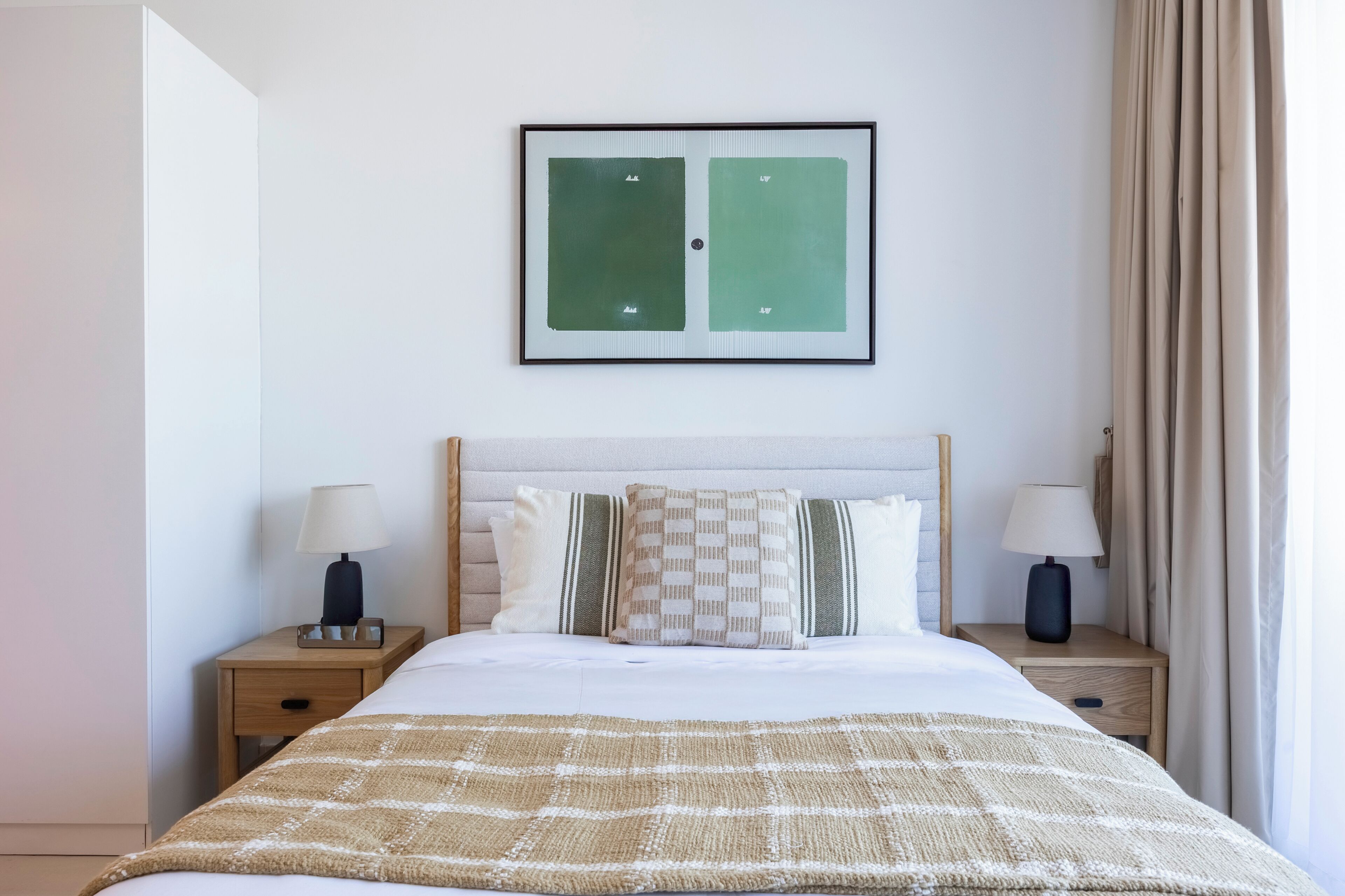 Habitación de hotel ordenada con cama, dos mesitas de noche con lámparas, un cuadro enmarcados encima de la cama y una ventana con cortinas.