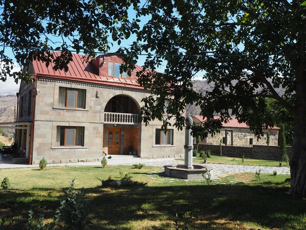 A stone building with a red roof, a central entrance with a wooden door, and a small garden area with a fountain.