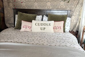 Una cama con un edredón estampado con motivos florales, un cabecero de madera oscura y varias almohadas con las palabras 'Cuddle Up' y 'Love'.