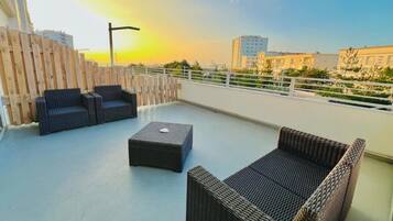 Una terraza en la azotea con muebles de mimbre, una cerca de madera y vistas a edificios al fondo.