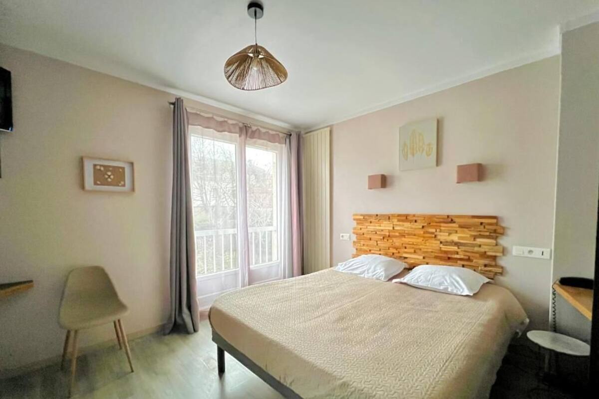 Une chambre à coucher comprenant un lit, une chaise, une table de chevet, un luminaire suspendu et une fenêtre avec des rideaux.
