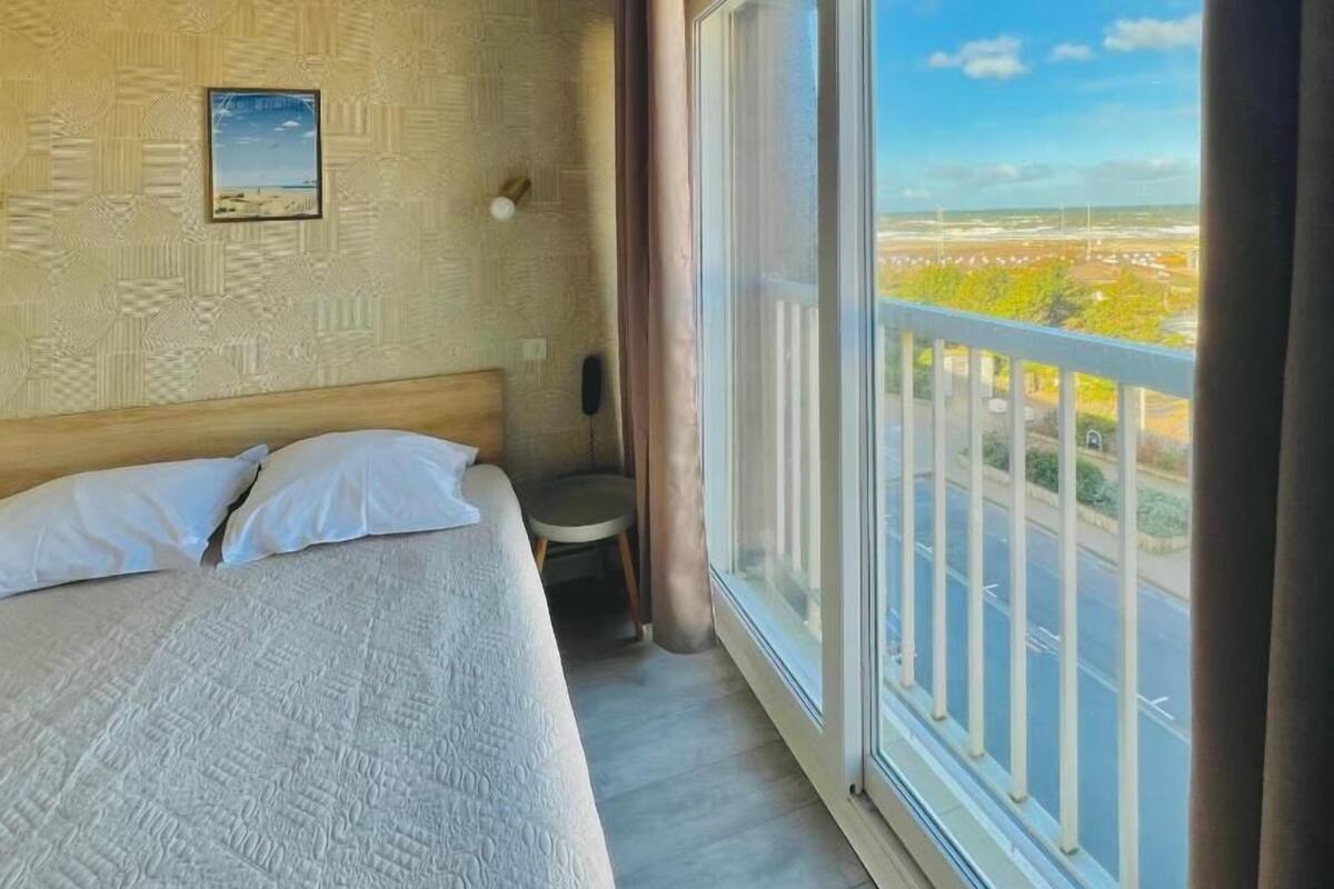 Une chambre d’hôtel avec un grand lit, une petite table, une chaise, une fenêtre donnant sur un parking et sur de la verdure, et un mur décoré d’un motif.