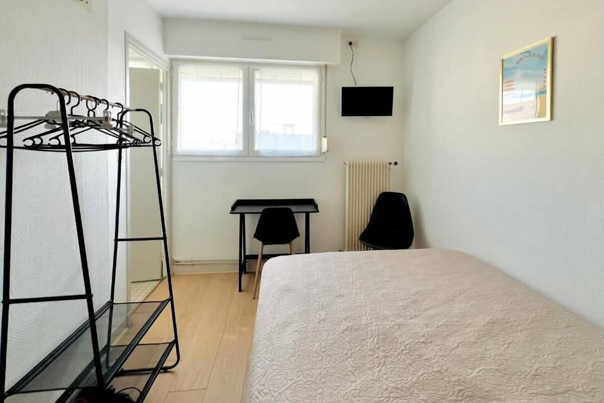 Une chambre à coucher avec un lit, un étendoir à linge en métal noir, un bureau avec une chaise et un radiateur.