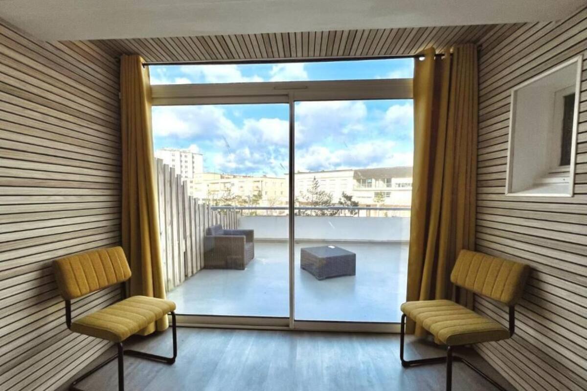 Une chambre moderne dotée d’une grande porte-fenêtre coulissante donnant sur un balcon extérieur, de deux chaises jaunes et d’un tableau encadré accroché au mur.