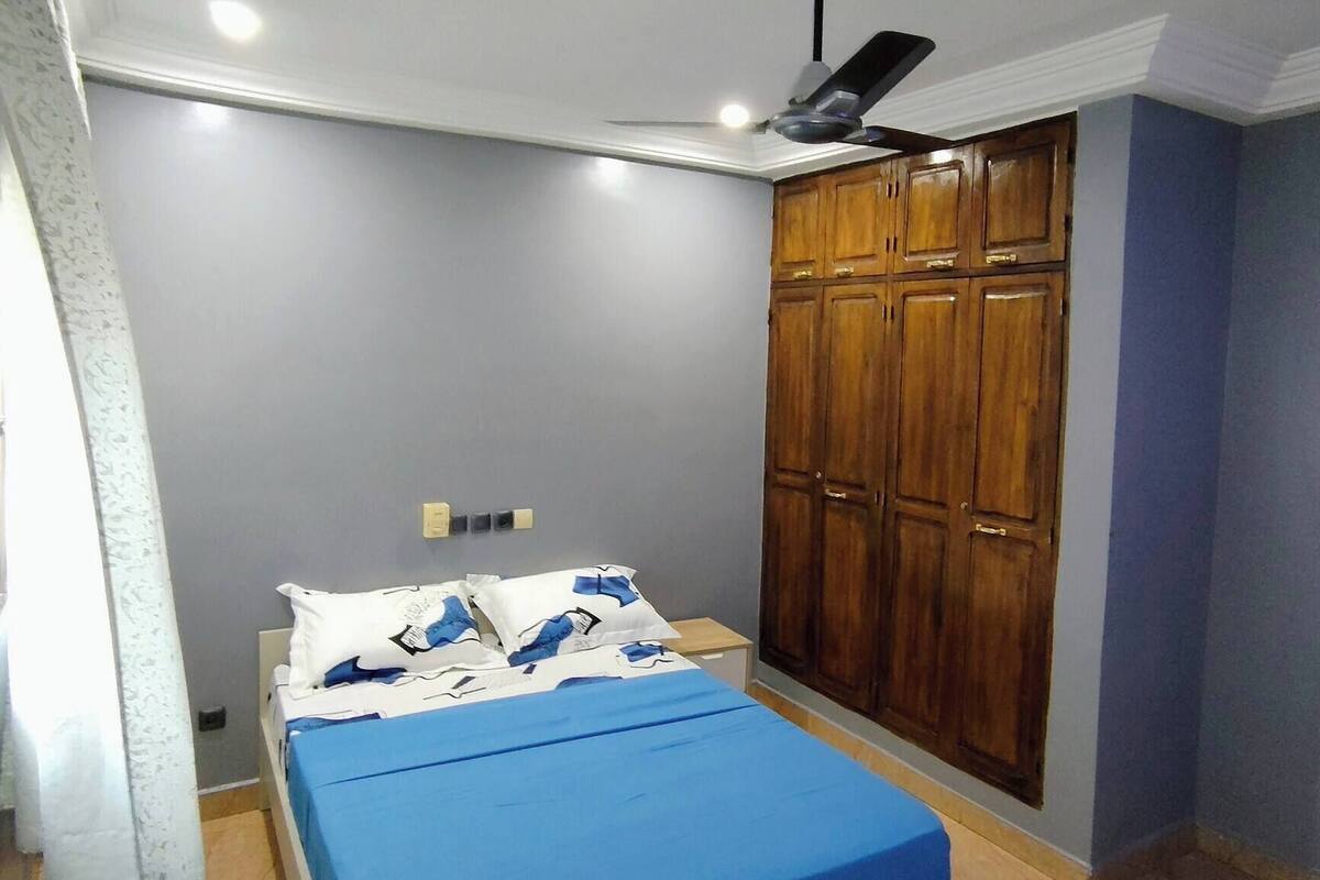 Une chambre à coucher avec un lit, un ventilateur de plafond, une armoire et une fenêtre avec des barreaux.