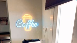 Een keukenhoek met een koffieapparaat, een waterkoker en twee kopjes op een houten tafel, met een neon bord met de tekst 'coffee' aan de muur.
