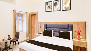 Una habitación de hotel con cama, escritorio, silla, mesita de noche, un florero con flores y tres cuadros en la pared.