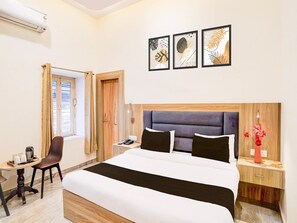 A hotel room with a bed, a desk, a chair, a nightstand, a vase with flowers, and three framed pictures on the wall. - Deluxe Hotel Shivam Residency (Dehradun)