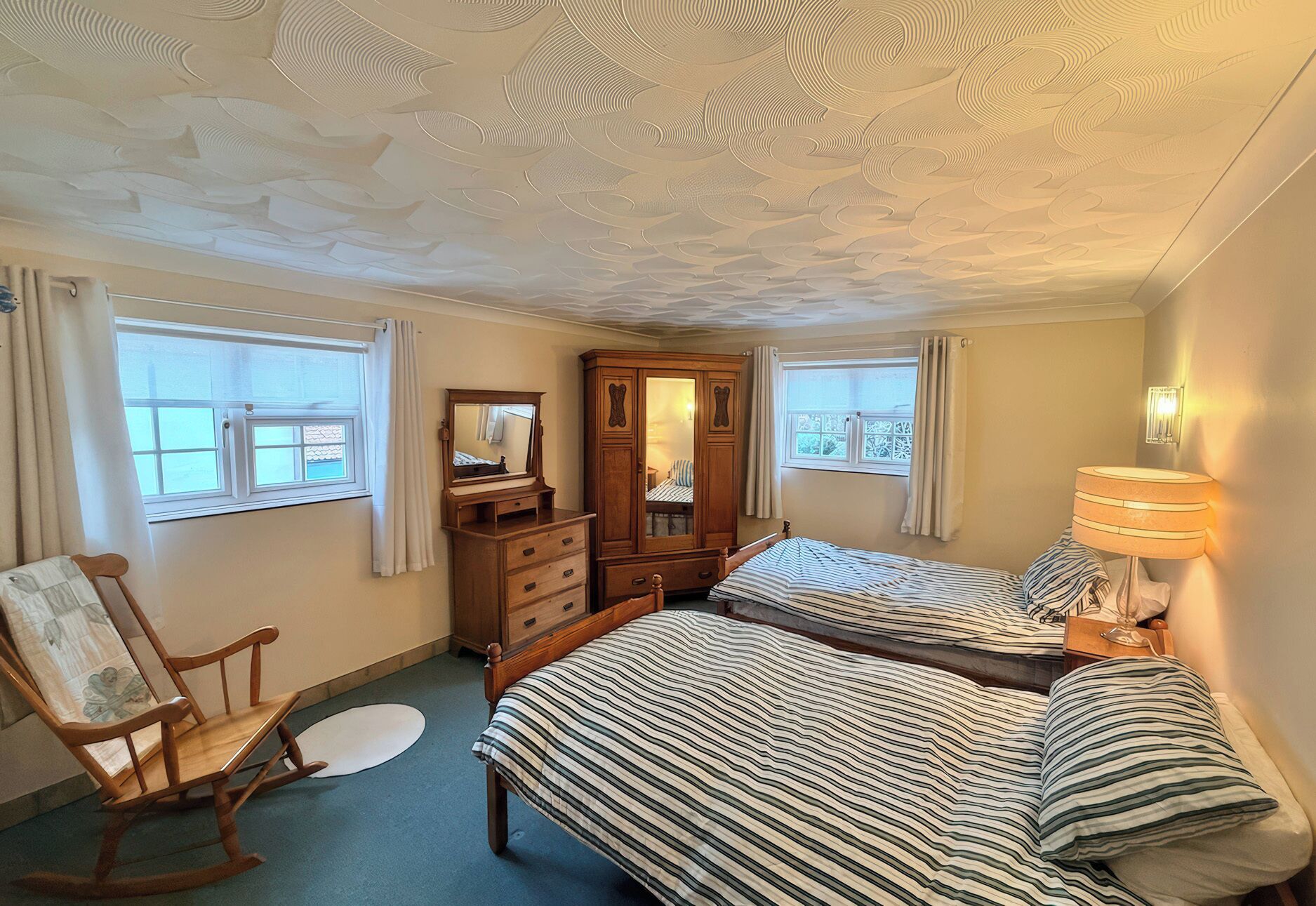 A bedroom with two beds, a wooden wardrobe, a rocking chair, and a window with curtains.